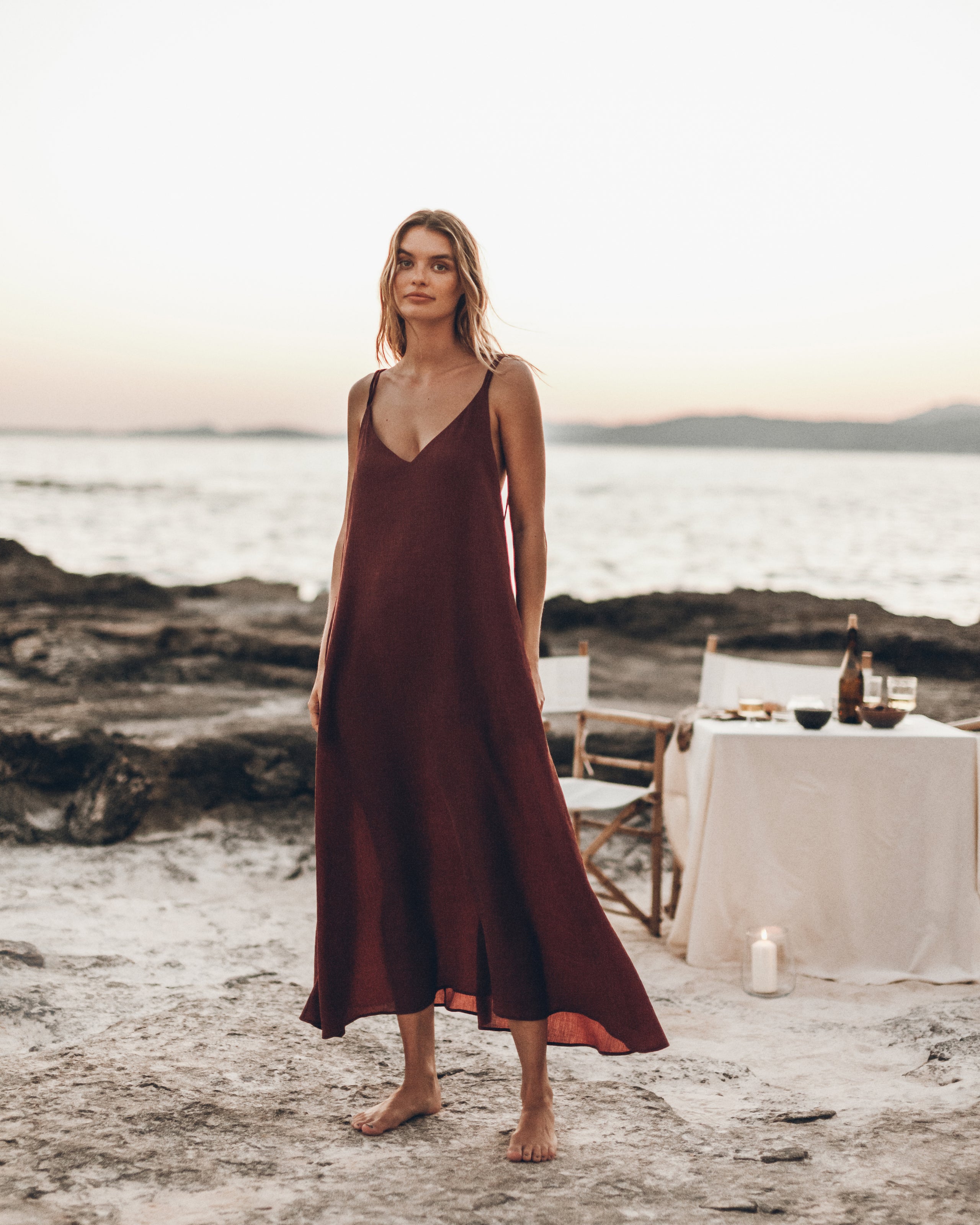 Woman in a burgundy dress standing on a rocky beach with a table set in the background.