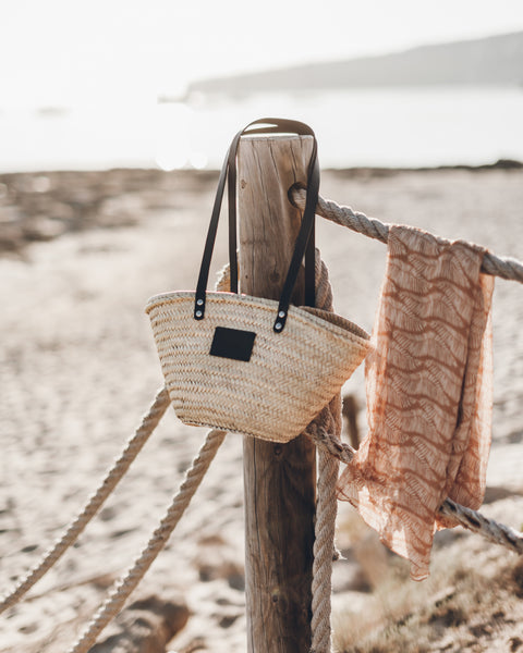 Woven bag with a black tag hanging on a wooden post by the beach.