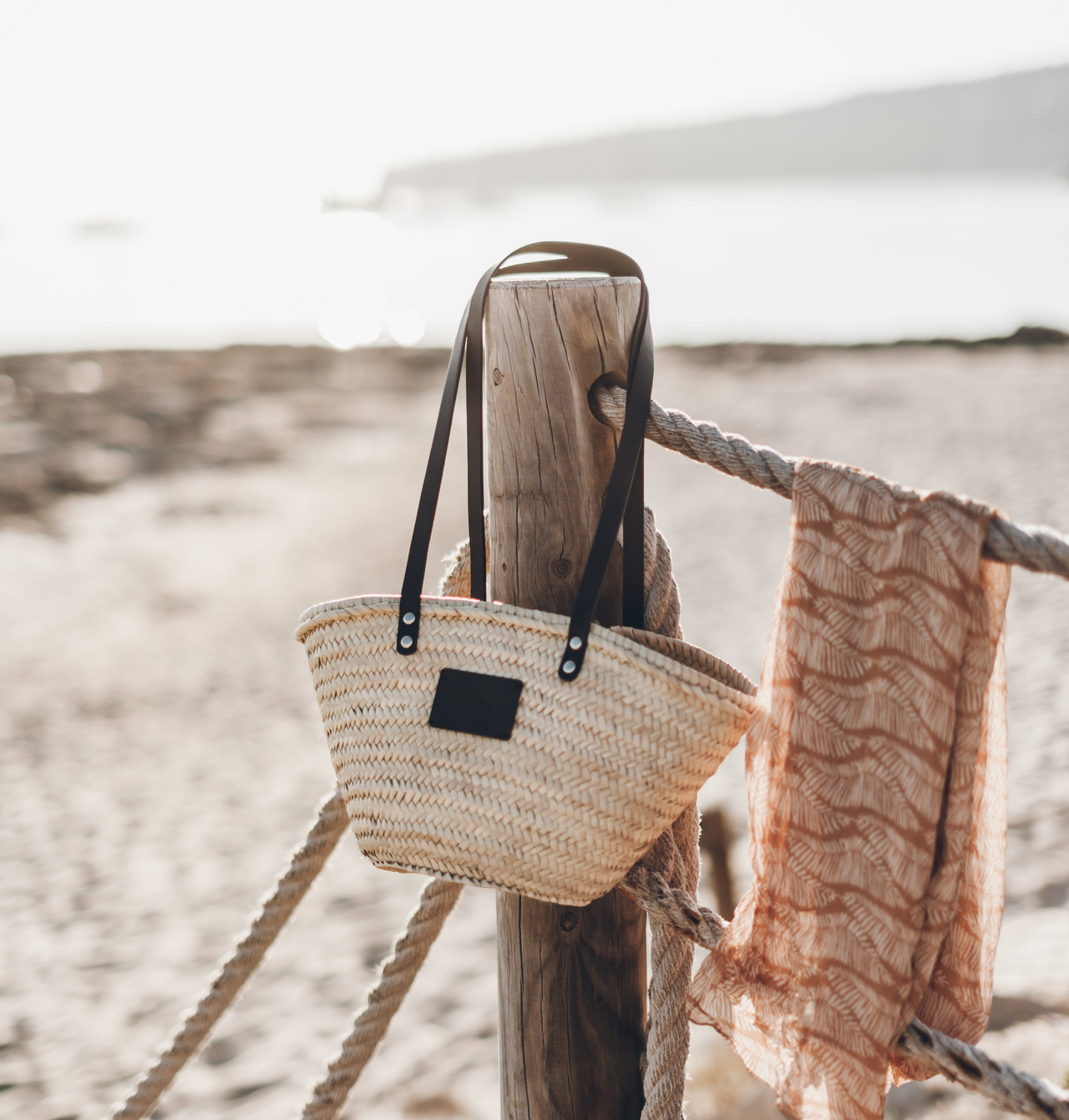 Woven bag and towel hanging on a wooden post with a beach background