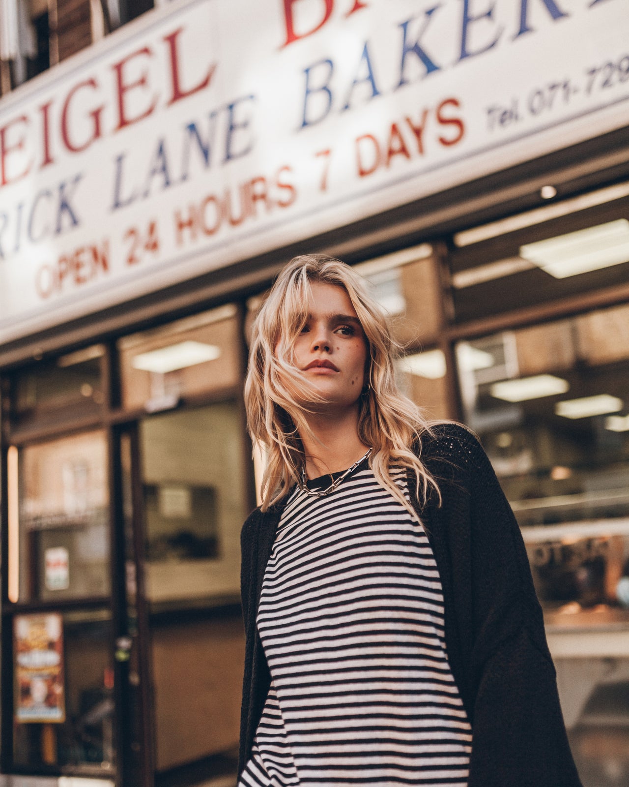 A blonde woman wearing a black and white striped top with a black cardigan stands in front of a bakery storefront.
