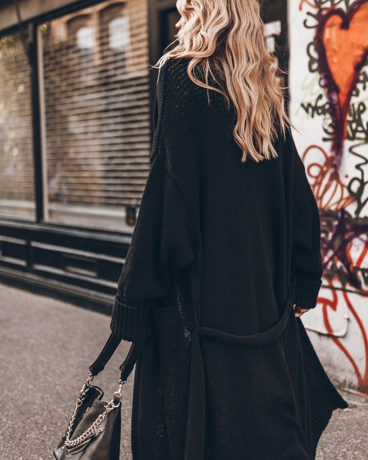 A woman from behind wearing a long black knit cardigan, carrying a black handbag with a silver chain strap, standing on a city street next to a graffiti-covered wall.