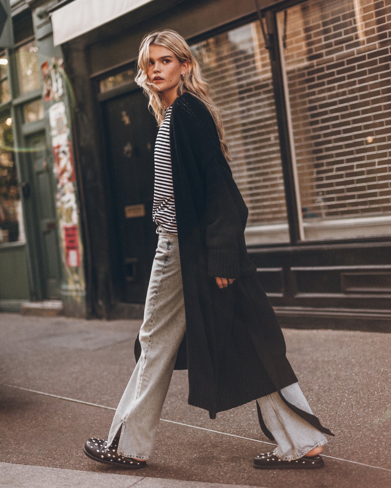 A woman walks on a city sidewalk wearing a long black open-front cardigan over a black-and-white striped top, light gray wide-leg jeans, and black studded mules.
