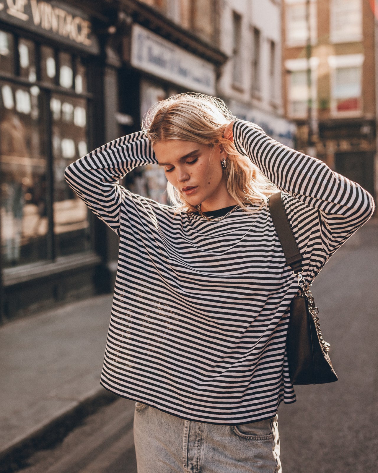 A woman posing on a city street wearing a black and white horizontally striped long-sleeve top, light-wash jeans, and carrying a black shoulder bag.