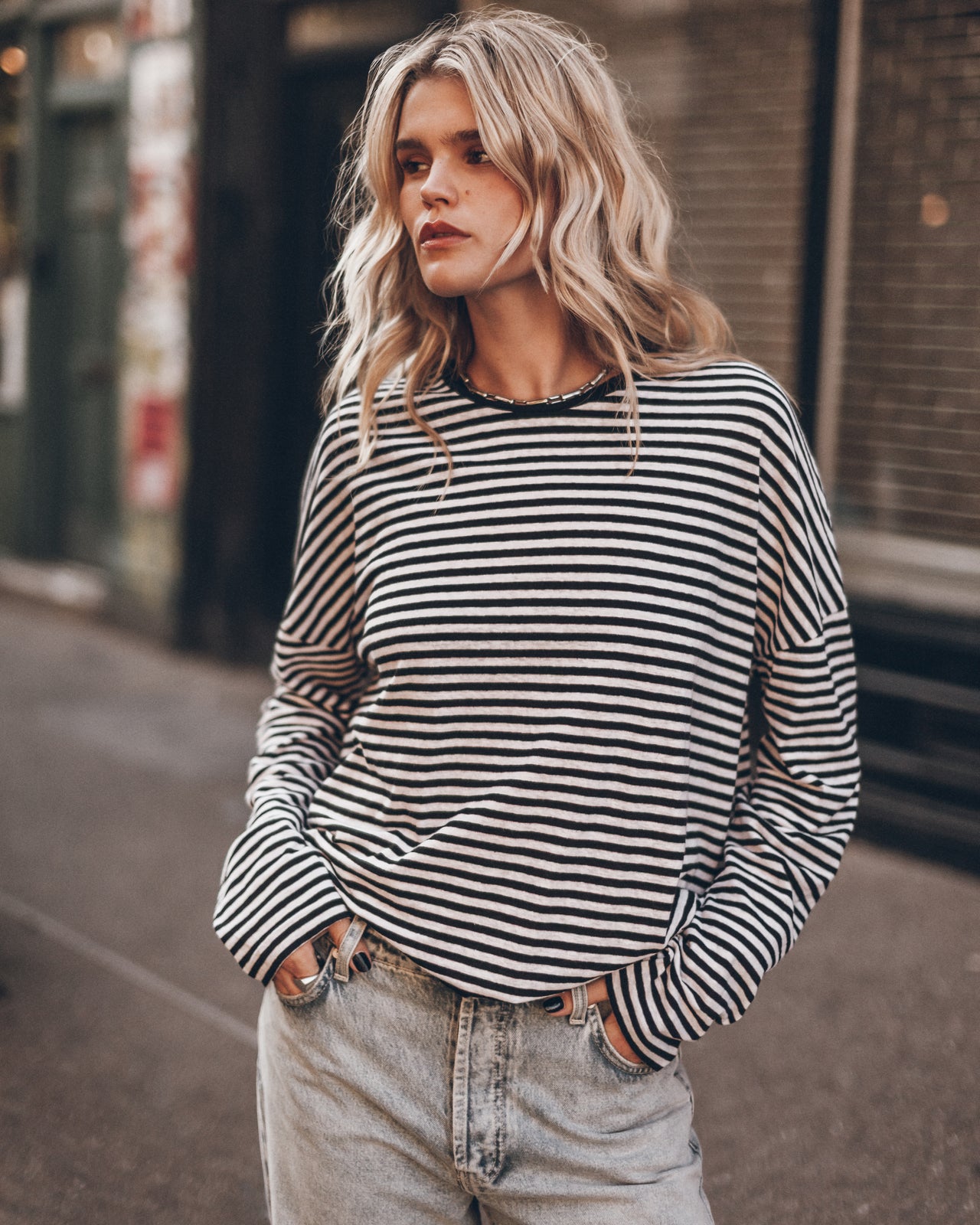 A woman wearing a black and white horizontally striped longsleeve  top with light-wash jeans, standing on an outdoor city street.