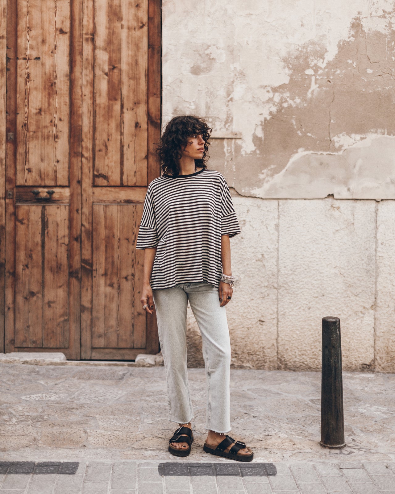 Woman standing on a street wearing a striped linen t-shirt, light jeans, and black leather sandals.