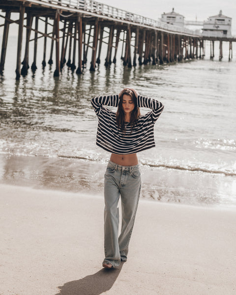 Woman in a striped sweater and jeans standing on a beach with a pier in the background