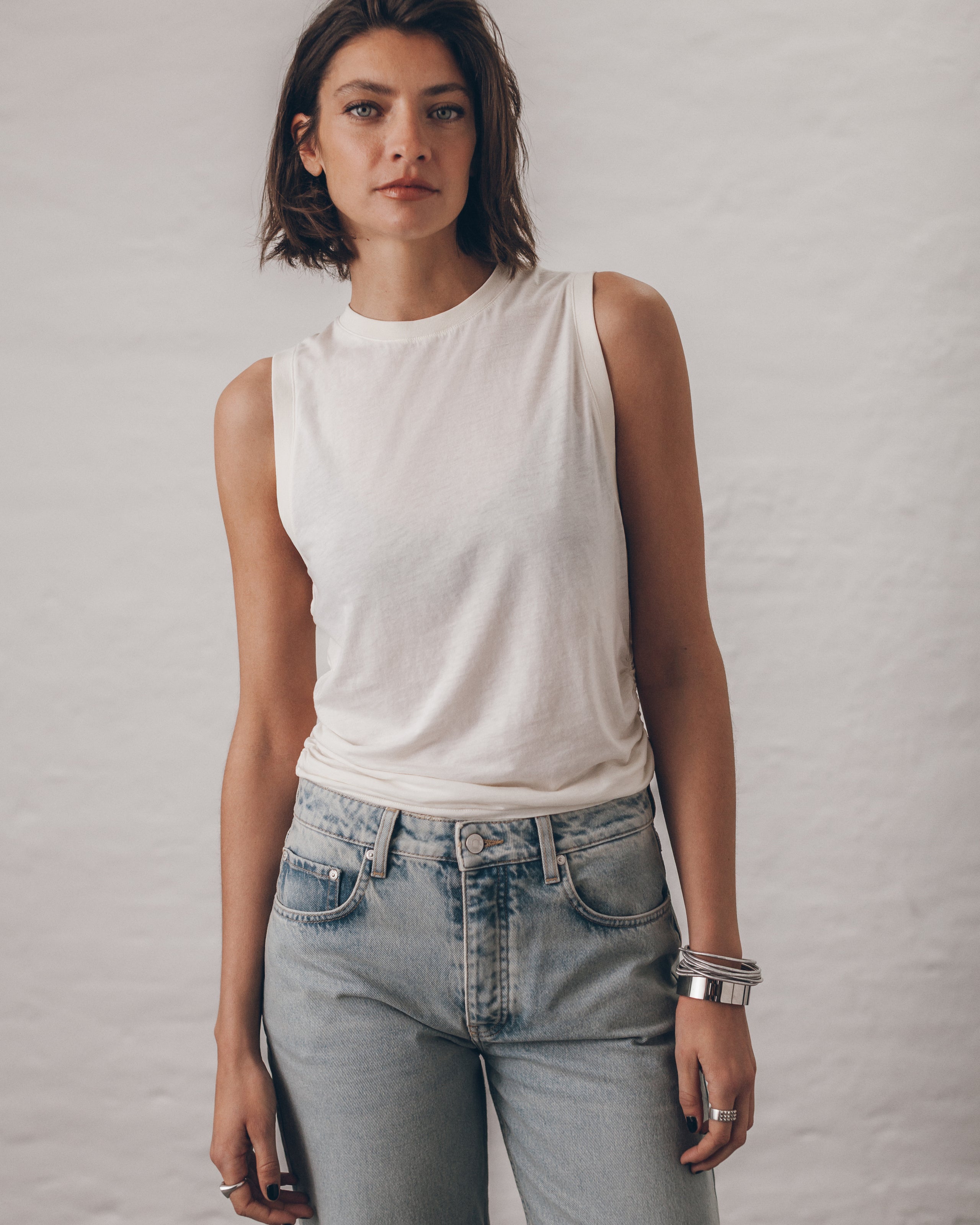 Woman wearing a white sleeveless top and light blue jeans against a plain background