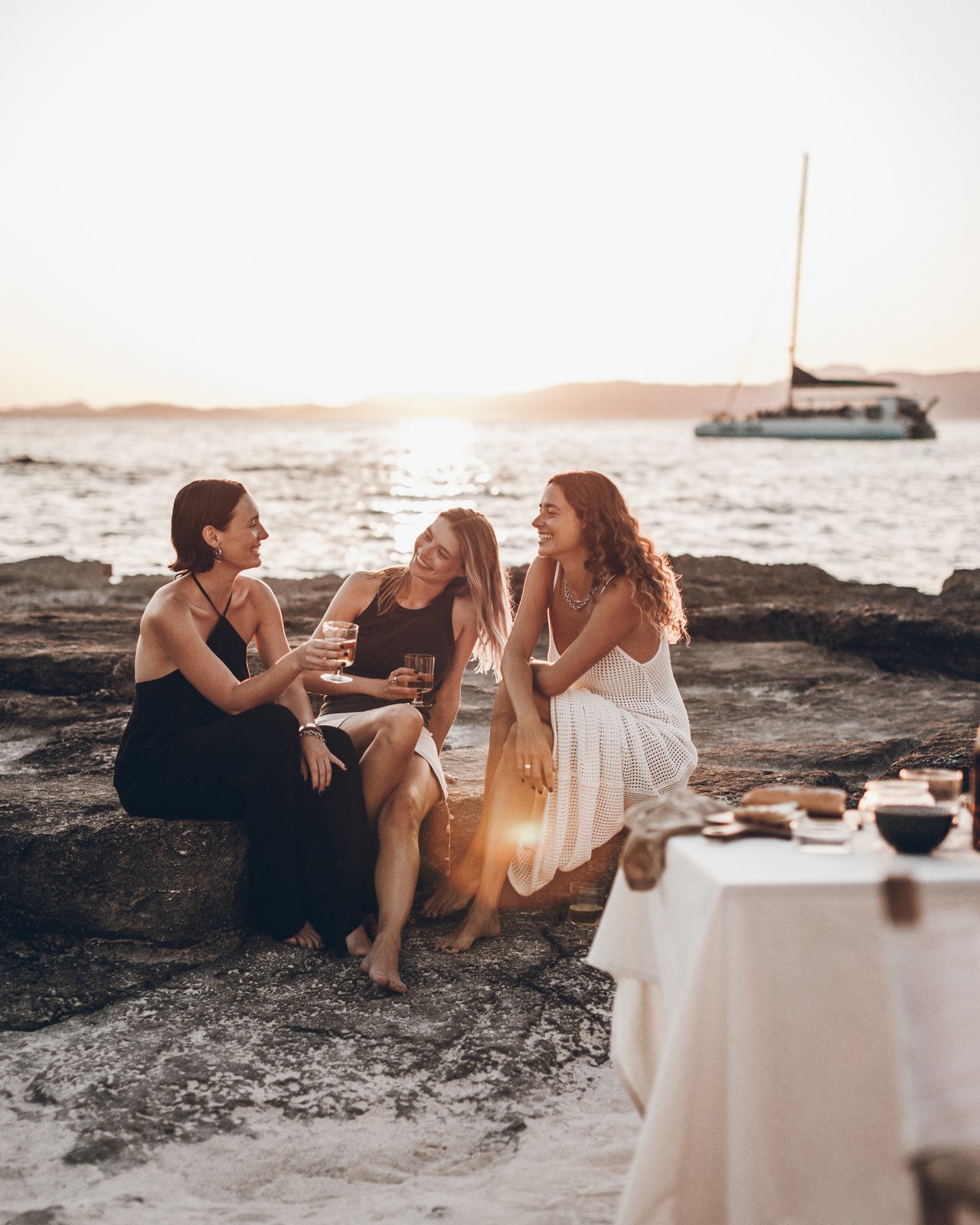 Three women sitting on rocks by the water with a sailboat in the background