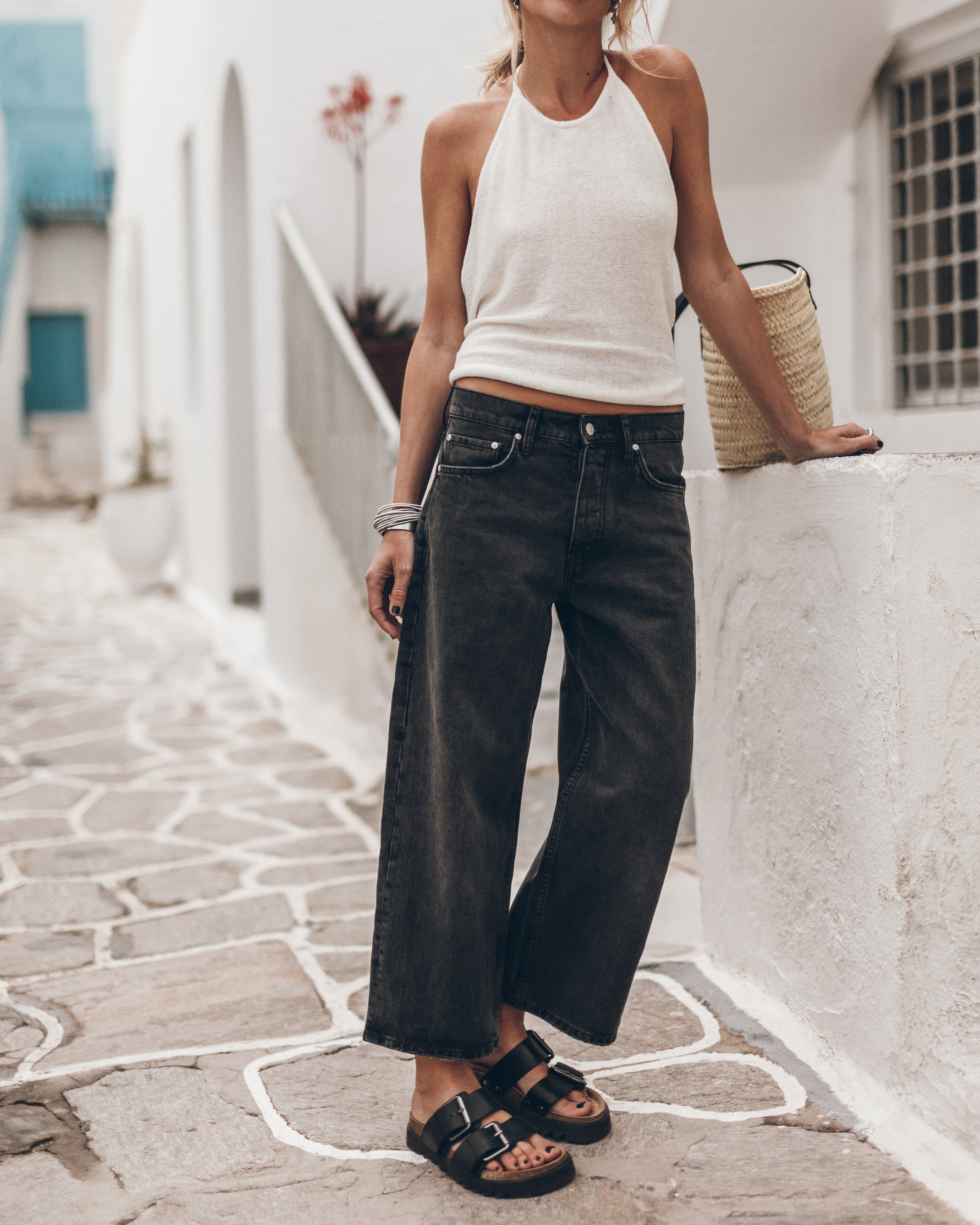 Woman wearing a white tank top and black jeans on a street, with a straw bag on her side.