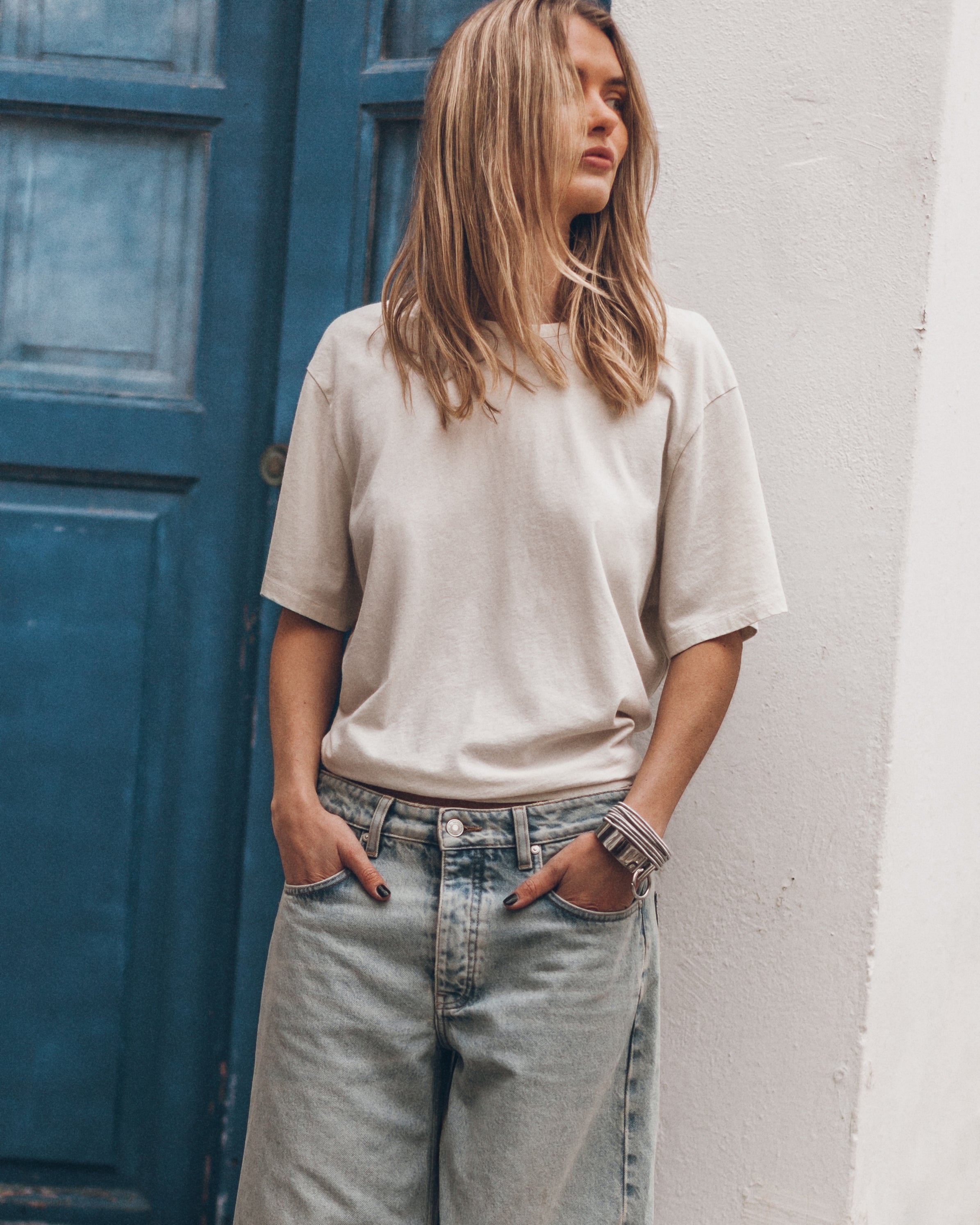 Woman wearing a light beige t-shirt and light blue jeans standing against a white wall with a blue door.