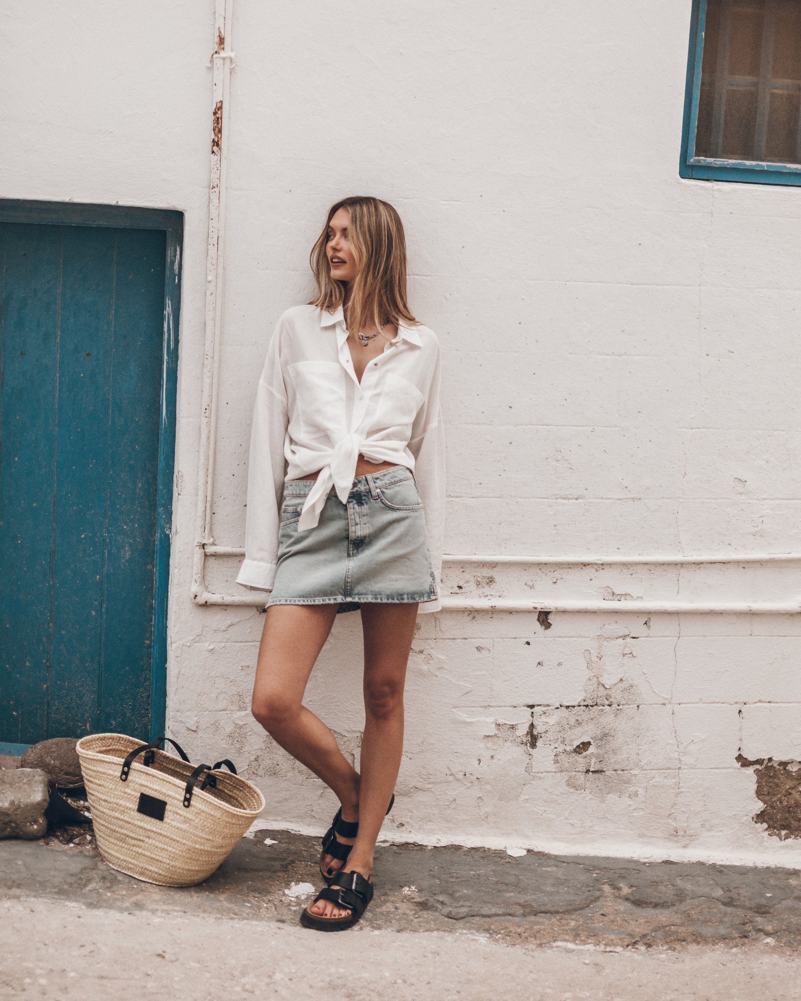 Woman in a white shirt and denim shorts standing against a white wall with a blue door.