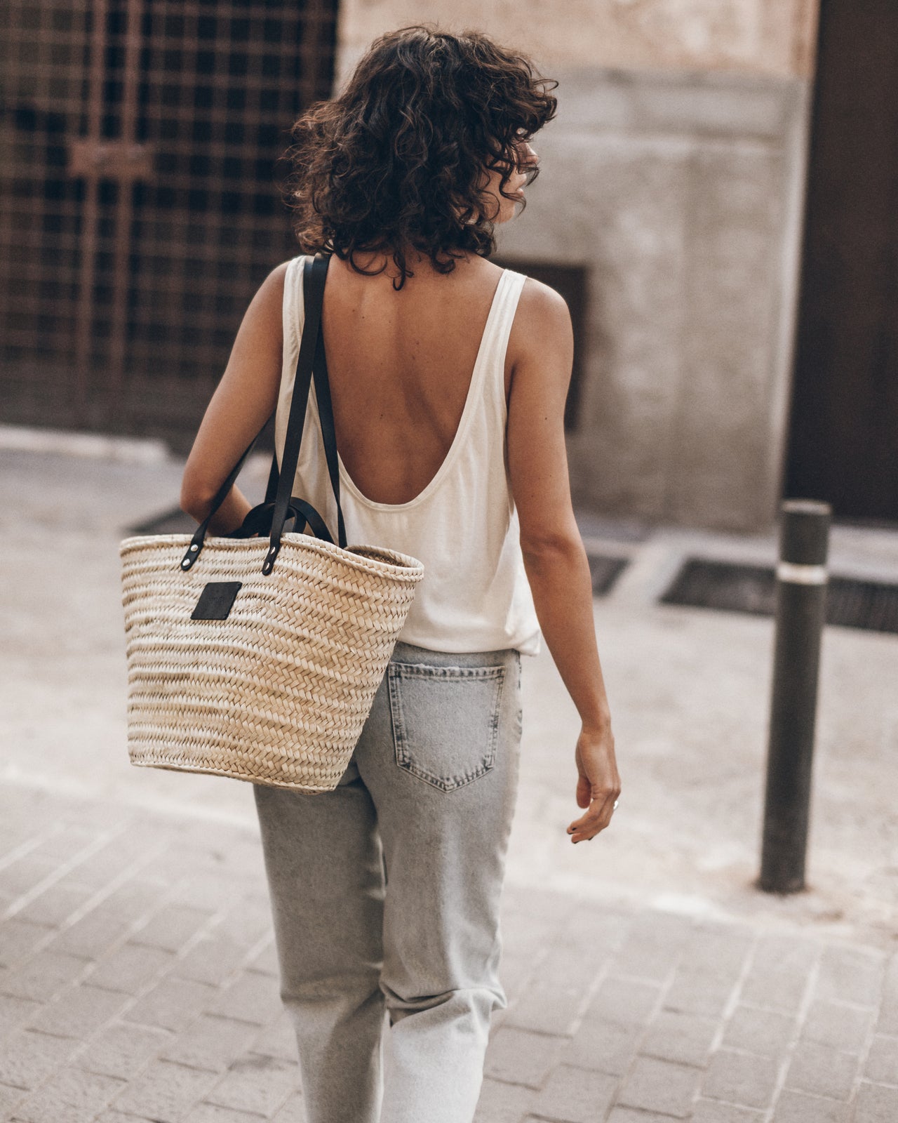 Woman walking on a street wearing a white top, light blue jeans, and a straw bag over one shoulder.