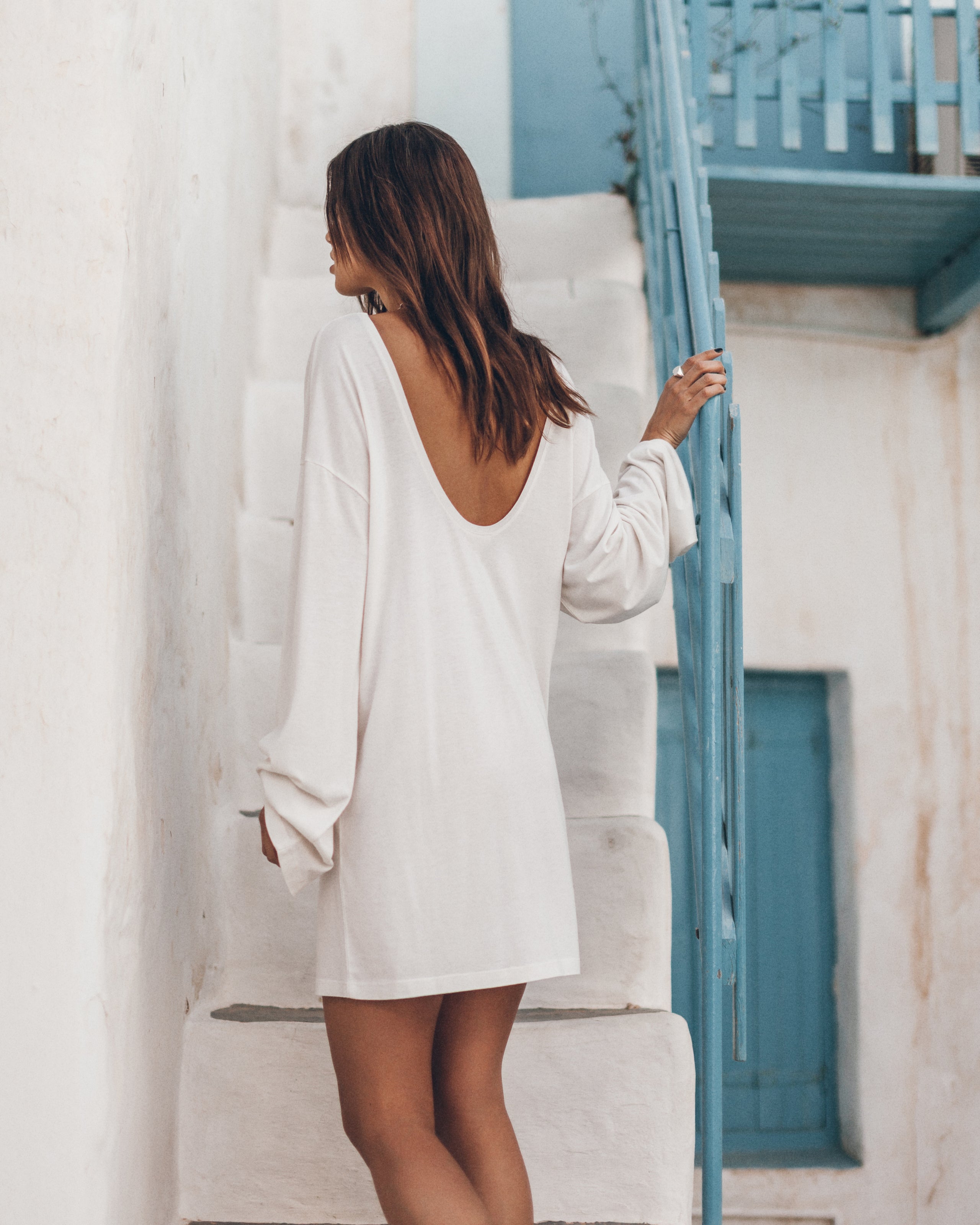 Woman in a white open-back dress standing in front of a white wall.