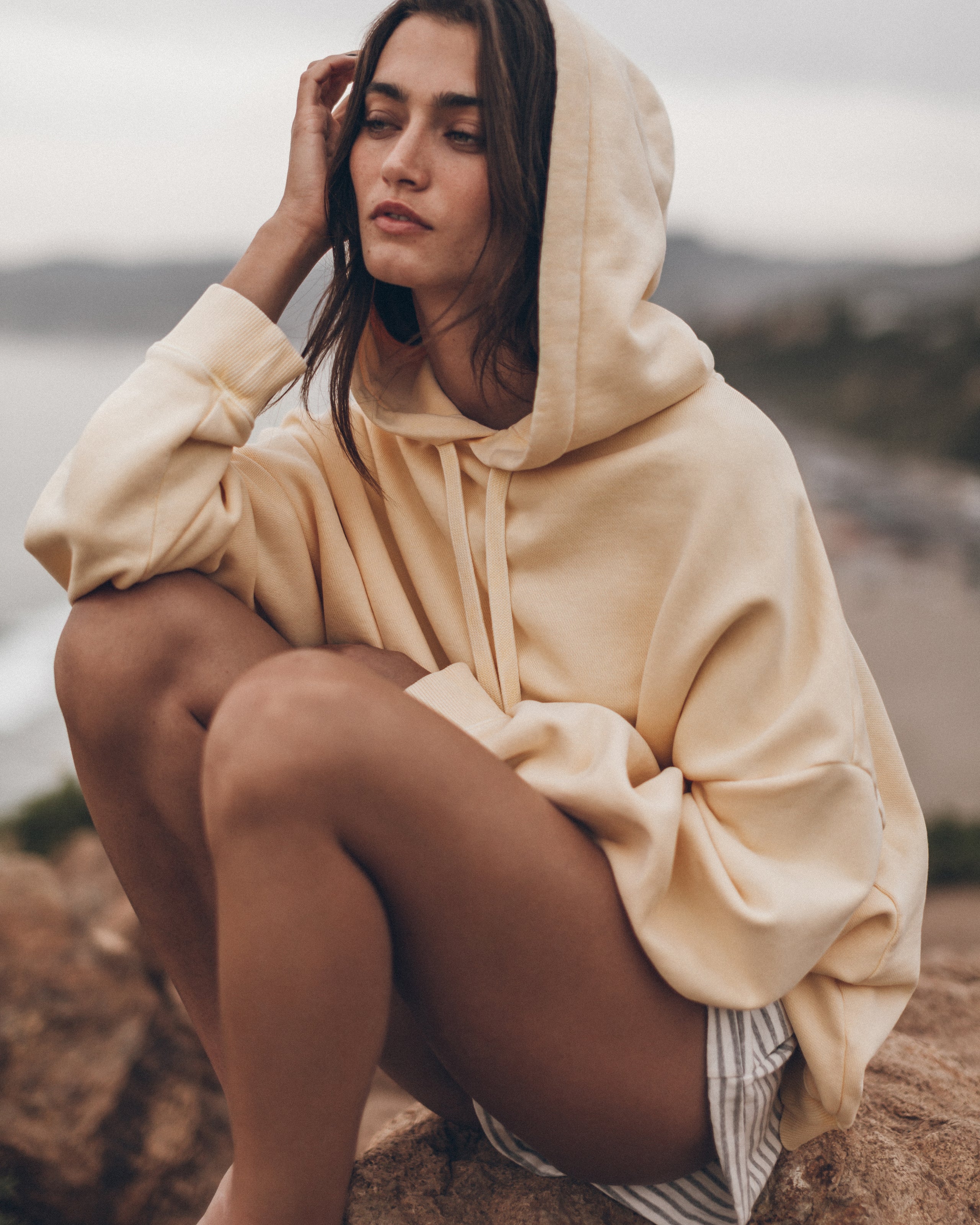 Woman wearing a beige hoodie sitting on a rock by a body of water.