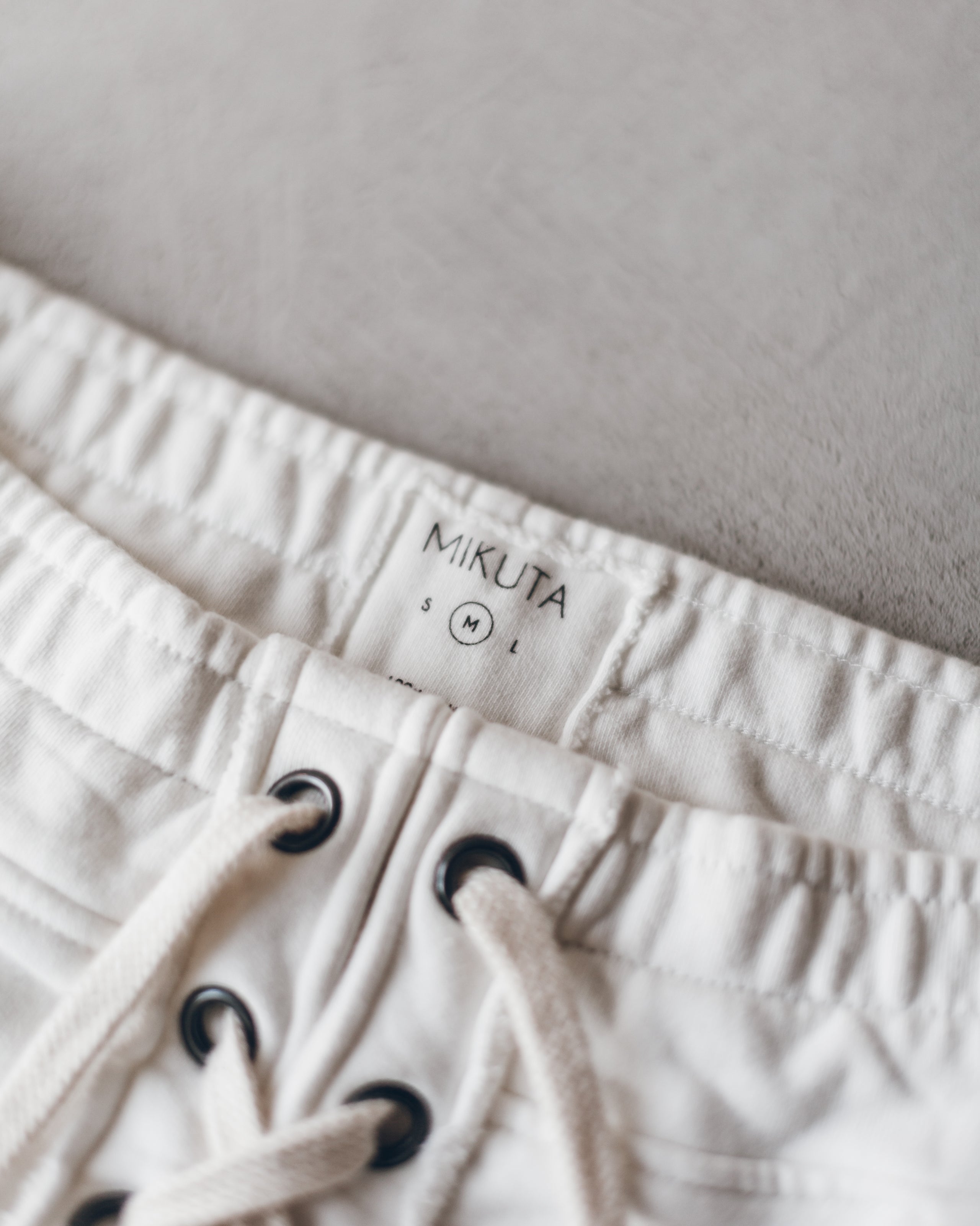 Ecru-colored casual shorts with elastic waistband and black grommet drawstring closure, featuring Mikuta label inside.