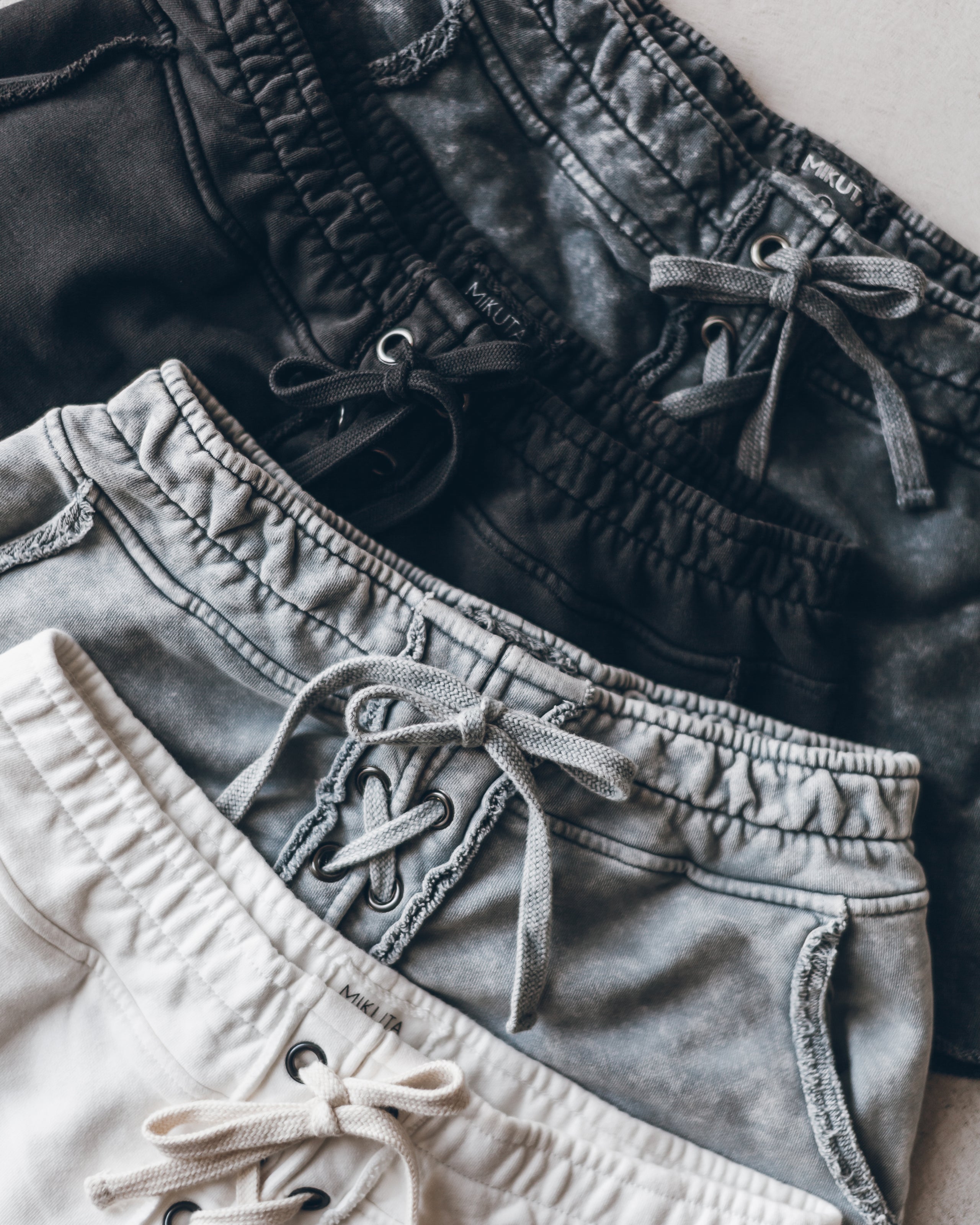 Three pairs of shorts stacked, featuring elastic waistbands with drawstrings in black, gray, and ecru colors.