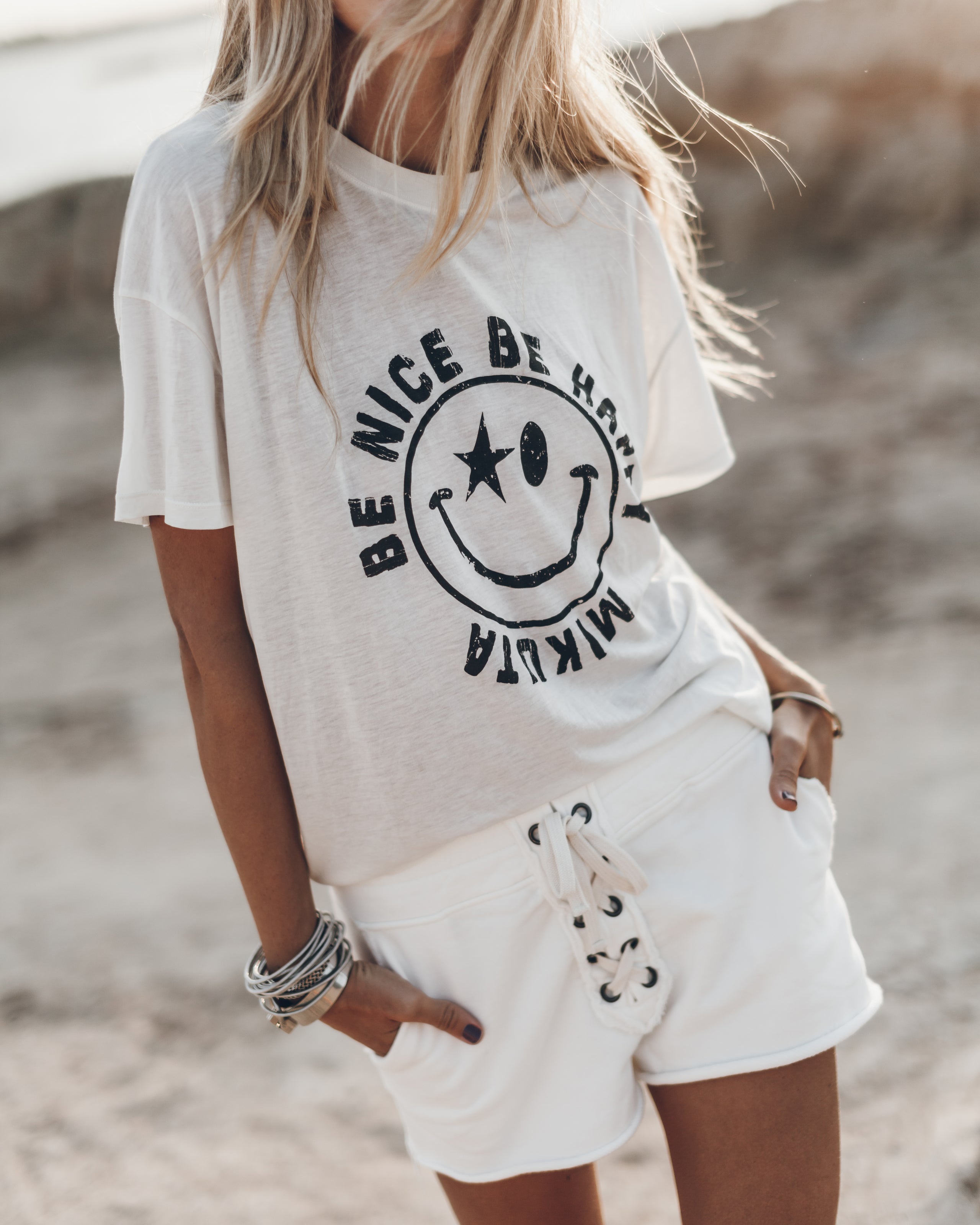 White oversized T-shirt with black smiley face and "Be Nice" text, paired with white lace-up shorts.