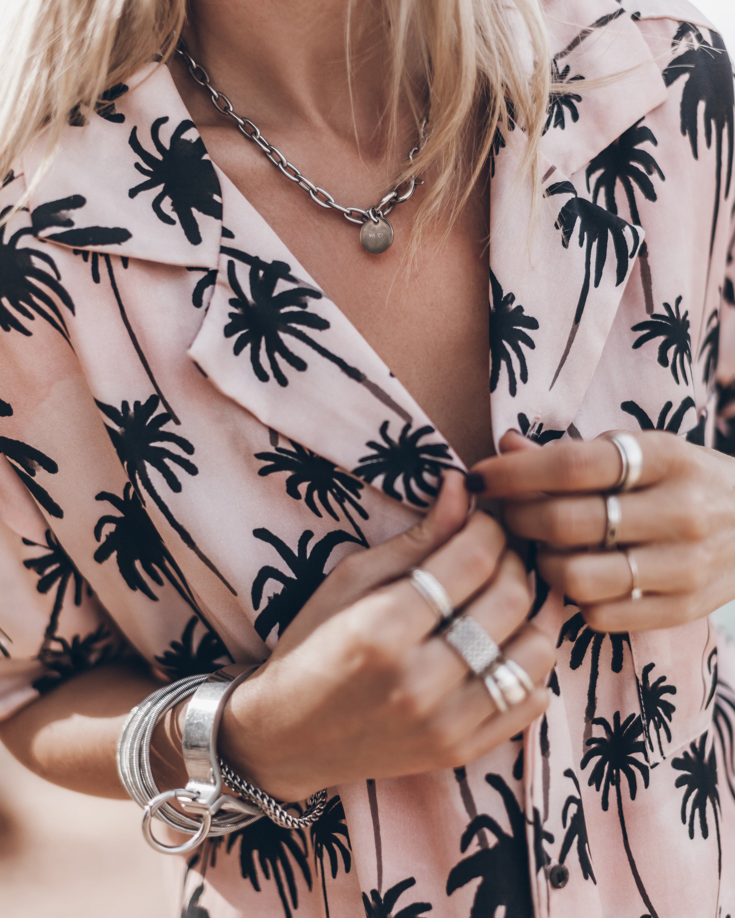 Light pink button-up shirt with black palm tree print and wide collar, styled with silver rings and bracelets.
